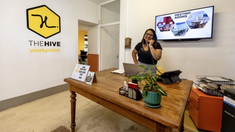A smiling woman talks on the phone behind a wooden desk in a reception area, with a large wall graphic displaying "THE HIVE workspace" logo and a screen advertising "MEETING ROOMS".