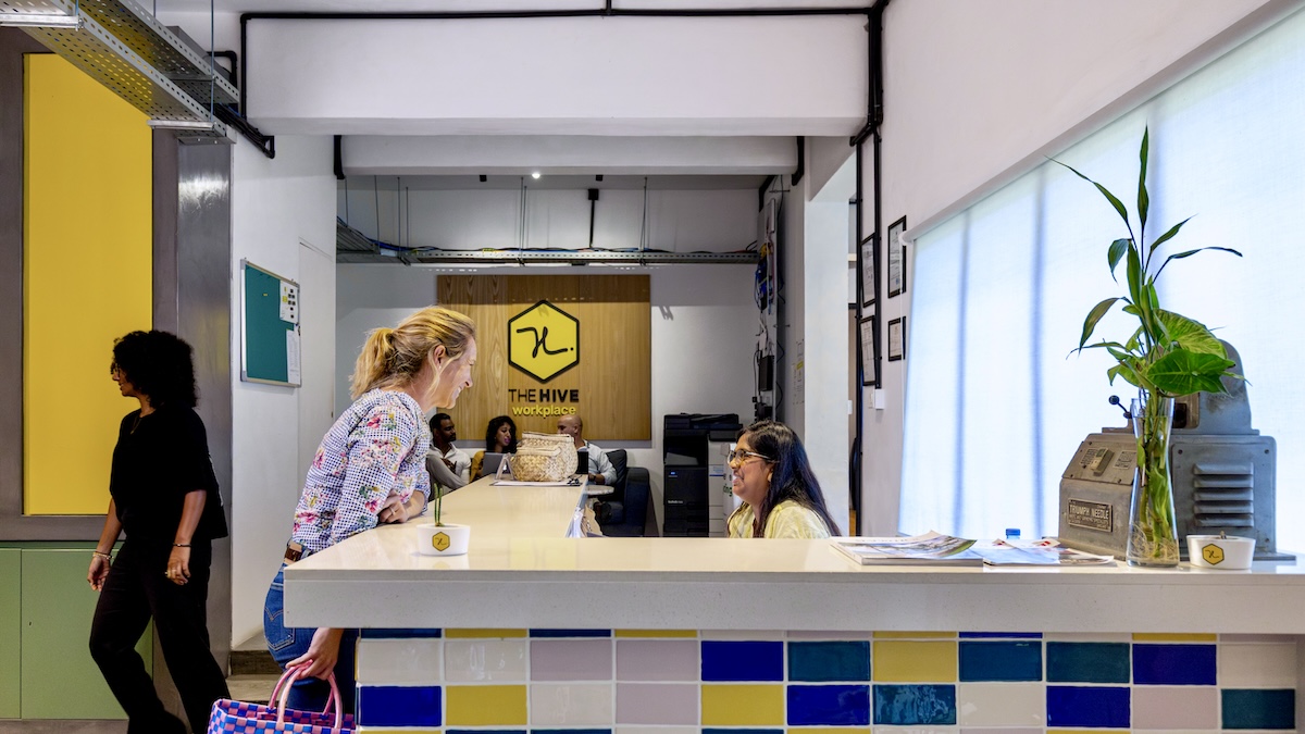 A bright, modern reception desk with yellow, blue, and white tiling. Two women are talking at the counter, and the wooden background clearly displays "The Hive" logo in a co-working workplace.