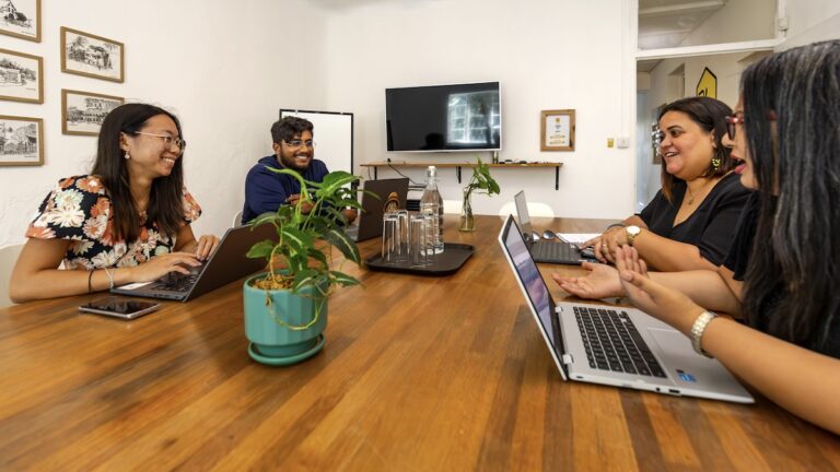 Four colleagues are seated around a long wooden table, smiling and collaborating with laptops in a bright hive workplace office setting, with a plant centerpiece.