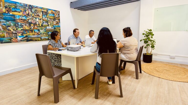 A small group of four people sits around a white rectangular table, collaborating in a bright, modern meeting room within the hive workplace space, with a large colorful collage on the wall.