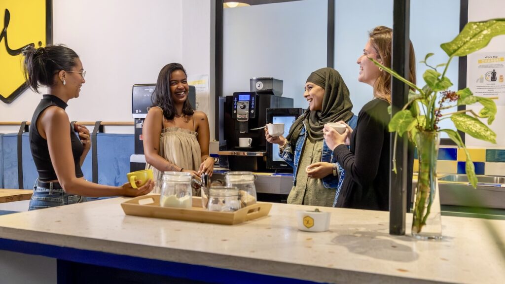 Four diverse women smiling and chatting while taking a coffee break near an espresso machine in the communal kitchen or break area of the hive co-working space.