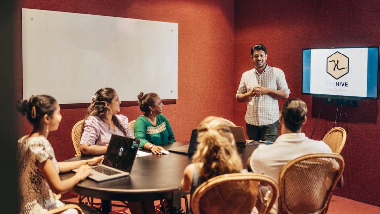 A man presents to a group of colleagues seated around a dark round table in a meeting room with red walls in the hive workplace; the screen displays "The Hive" logo.