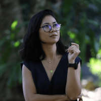 Headshot of a professional woman with dark hair and glasses, looking thoughtfully up and to the right, against a blurred green, natural background (suggesting a tranquil space outside the hive).