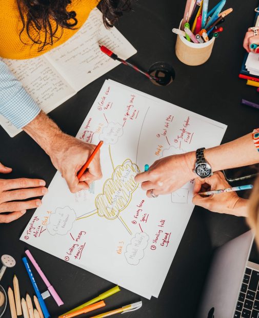 An overhead view of a group of people collaborating on a mind map drawn on a piece of paper, with hands, colorful pens, and a laptop visible on the black table in the hive.