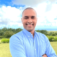 Headshot of a smiling man in a light blue shirt, with his arms crossed, standing outdoors in a lush, sunny landscape with the ocean visible in the distance (suggesting a break from the hive).