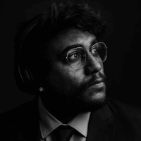 A dramatic black and white portrait of a man with glasses and a beard, wearing a suit and headphones, looking thoughtfully upwards, representing the creative energy within the hive.